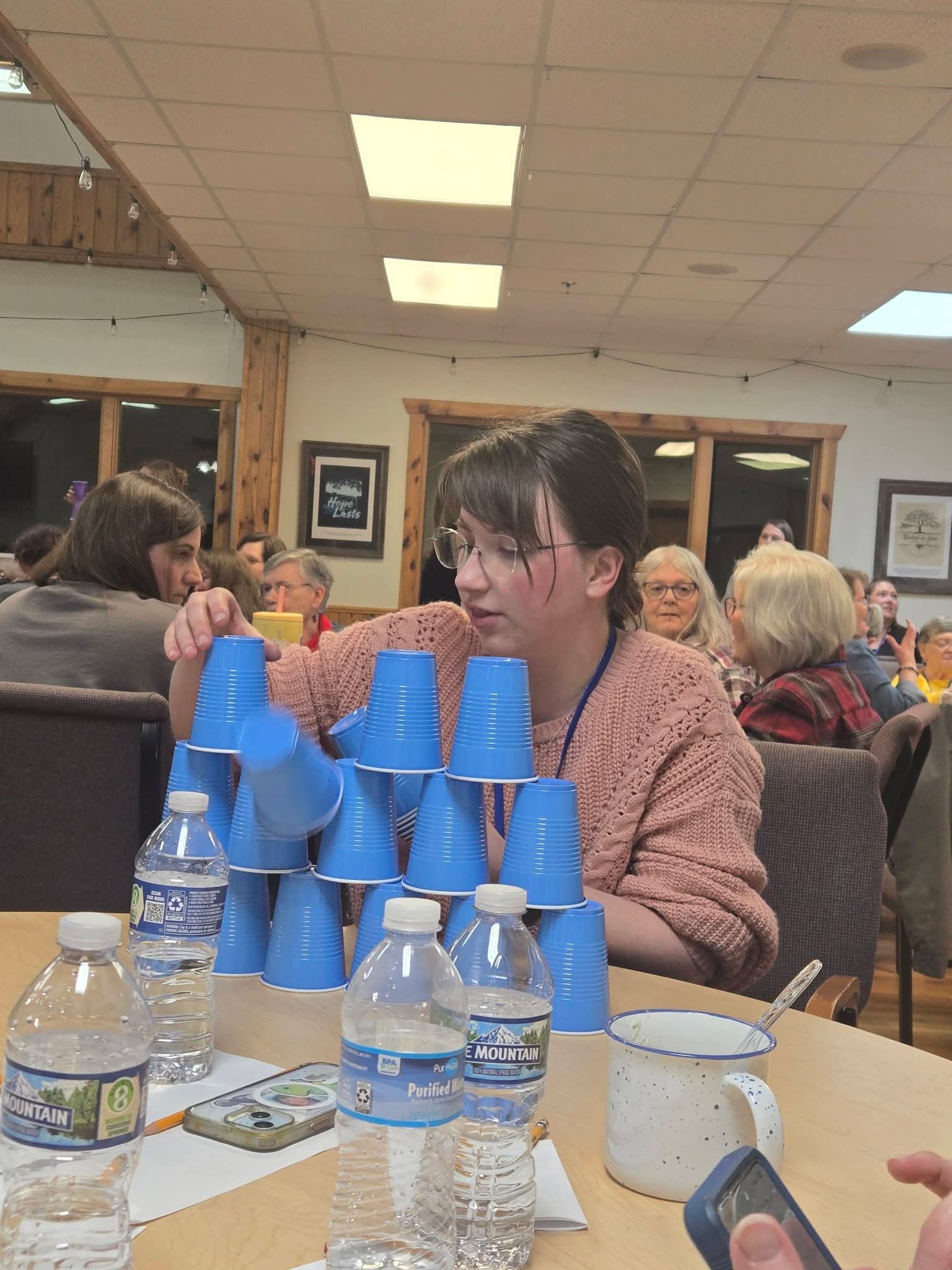 A group of people sitting at a table in a restaurant or cafe, with several water bottles, plastic cups stacked in a pyramid, a mug, and a smartphone on the table. The setting has wooden accents on the walls and framed pictures, with ceiling lights il