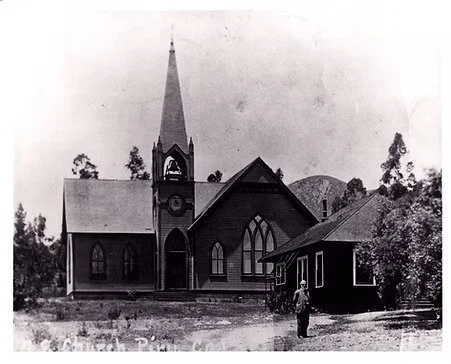 Black and white photo of a church with a tall steeple and a person standing in front of it.