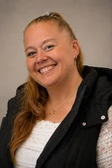 A smiling woman with long red hair wearing a black jacket, posing against a gray background.