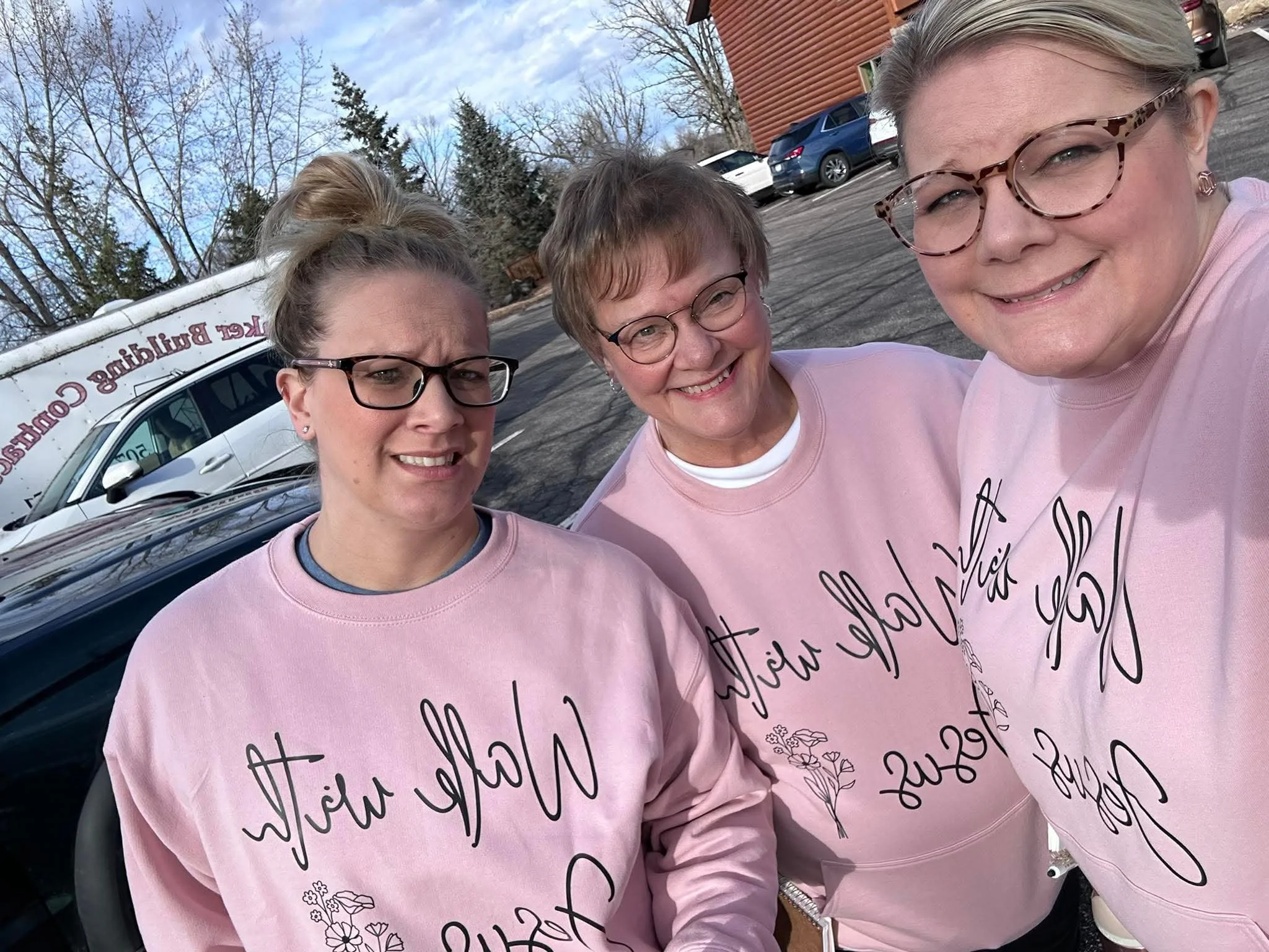 Three women taking a selfie outdoors, wearing pink sweatshirts with black text, in a parking lot with cars and trees in the background.