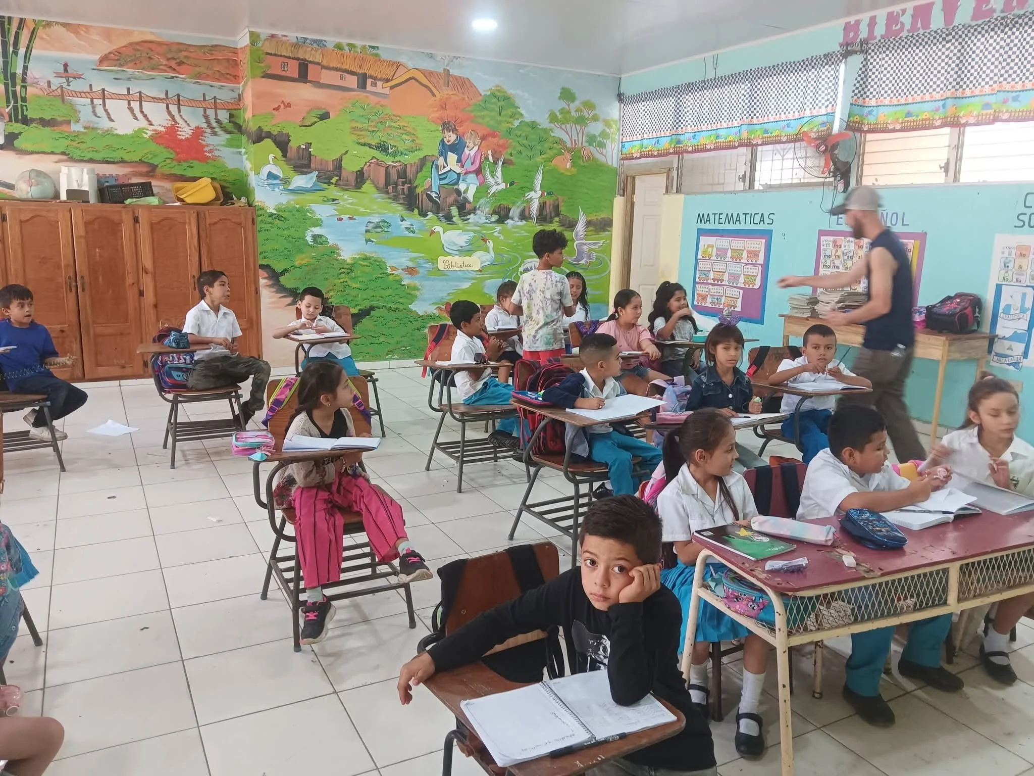 Classroom with students seated at desks and a colorful mural on the wall depicting nature scenes including lakes, trees, animals, and children reading.