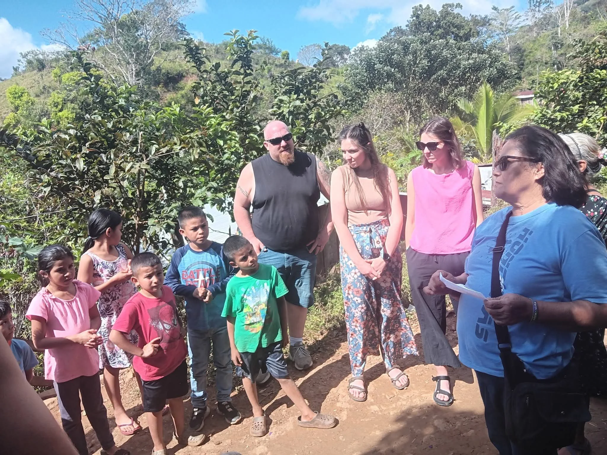 A group of children and adults standing outdoors in a rural, hilly area with green trees and blue sky. The adults are talking, and the children appear to be listening attentively.