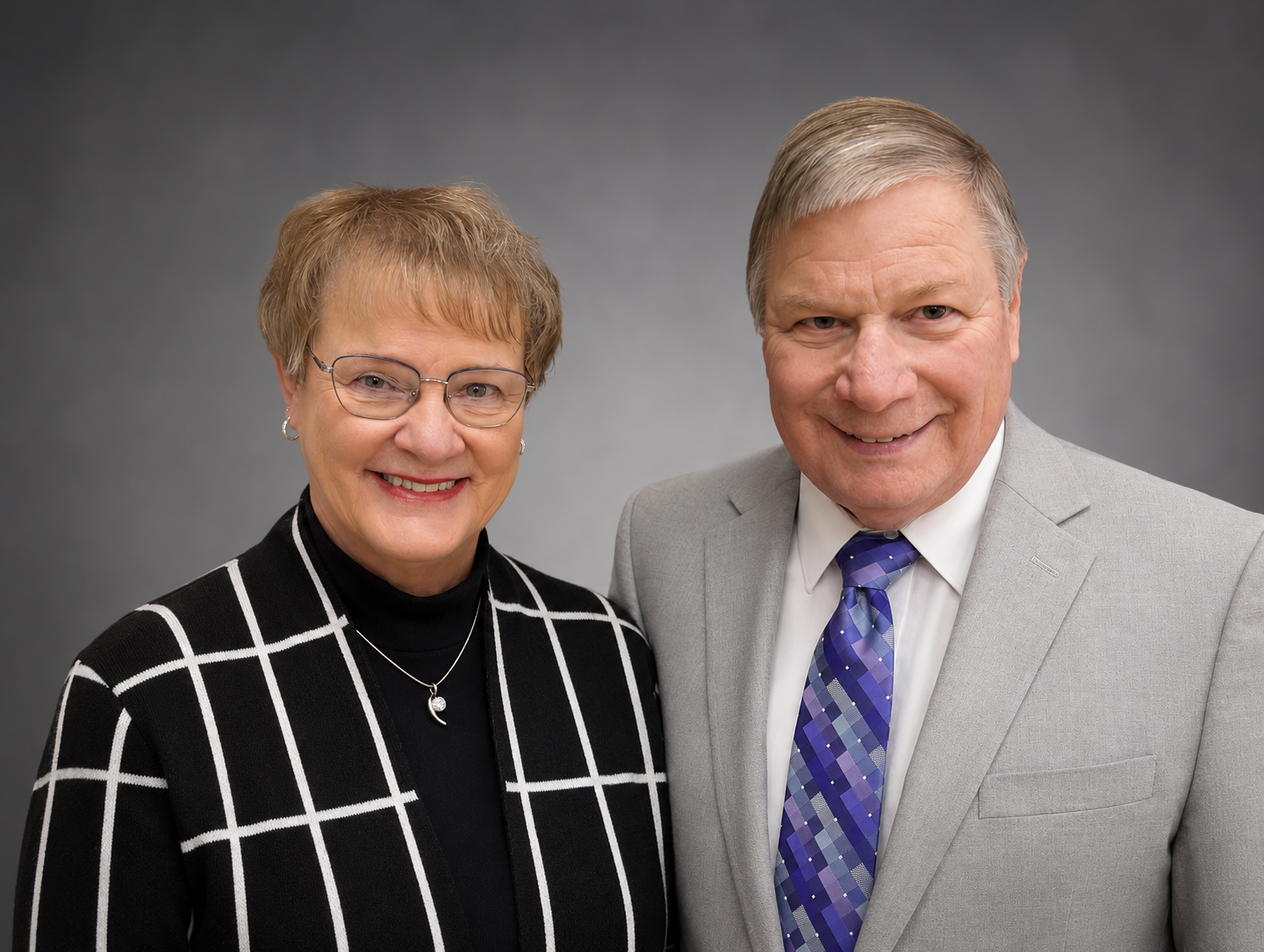 A smiling older woman with short hair, glasses, a black and white checkered blazer, and jewelry standing next to an older man with short gray hair, a light gray suit, white shirt, and blue checkered tie against a gray background.