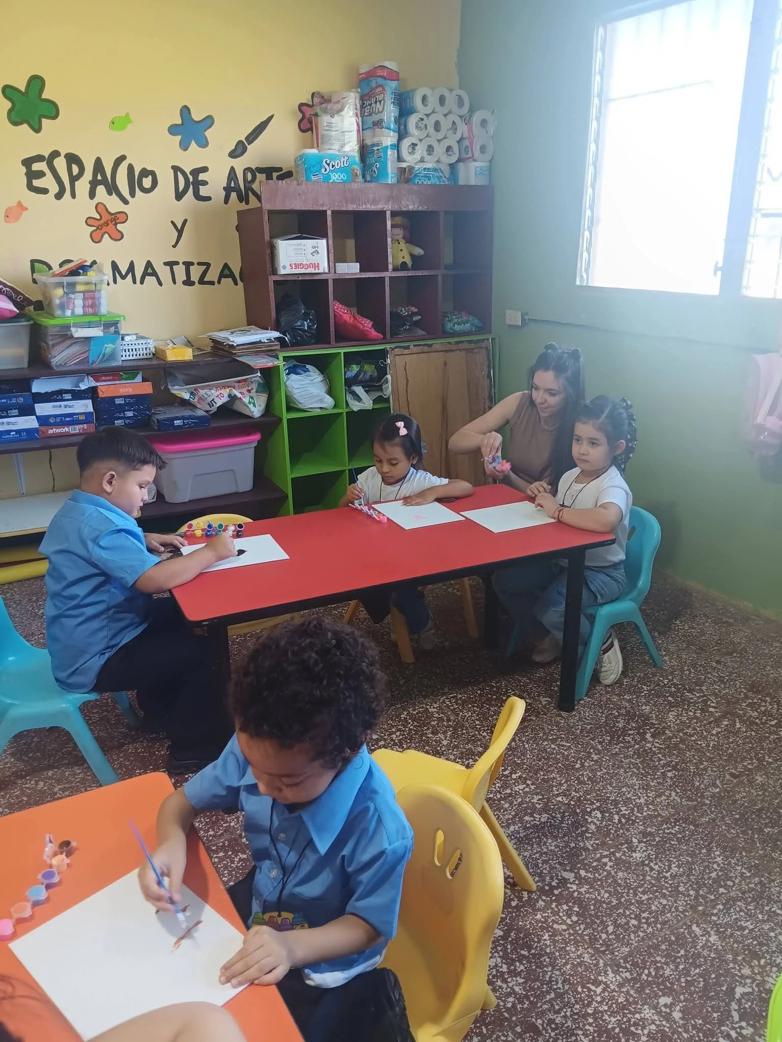 Children and a teacher engaged in arts and crafts activities at a table in a classroom with colorful walls and art supplies.
