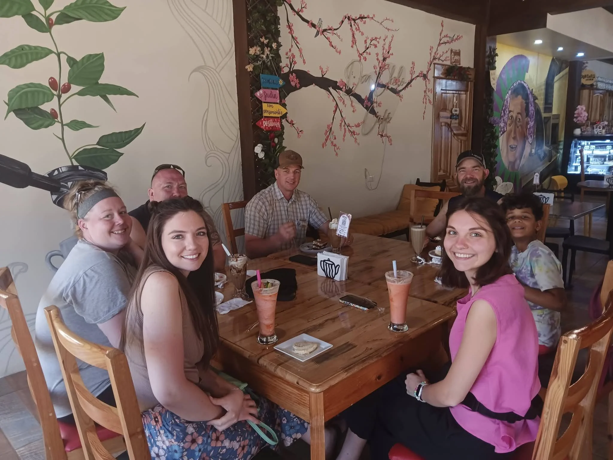 A group of people sitting around a wooden table in a restaurant, smiling at the camera with desserts and drinks in front of them. The background has wall decorations including floral murals and artwork, with a section of colorful wall art to the righ