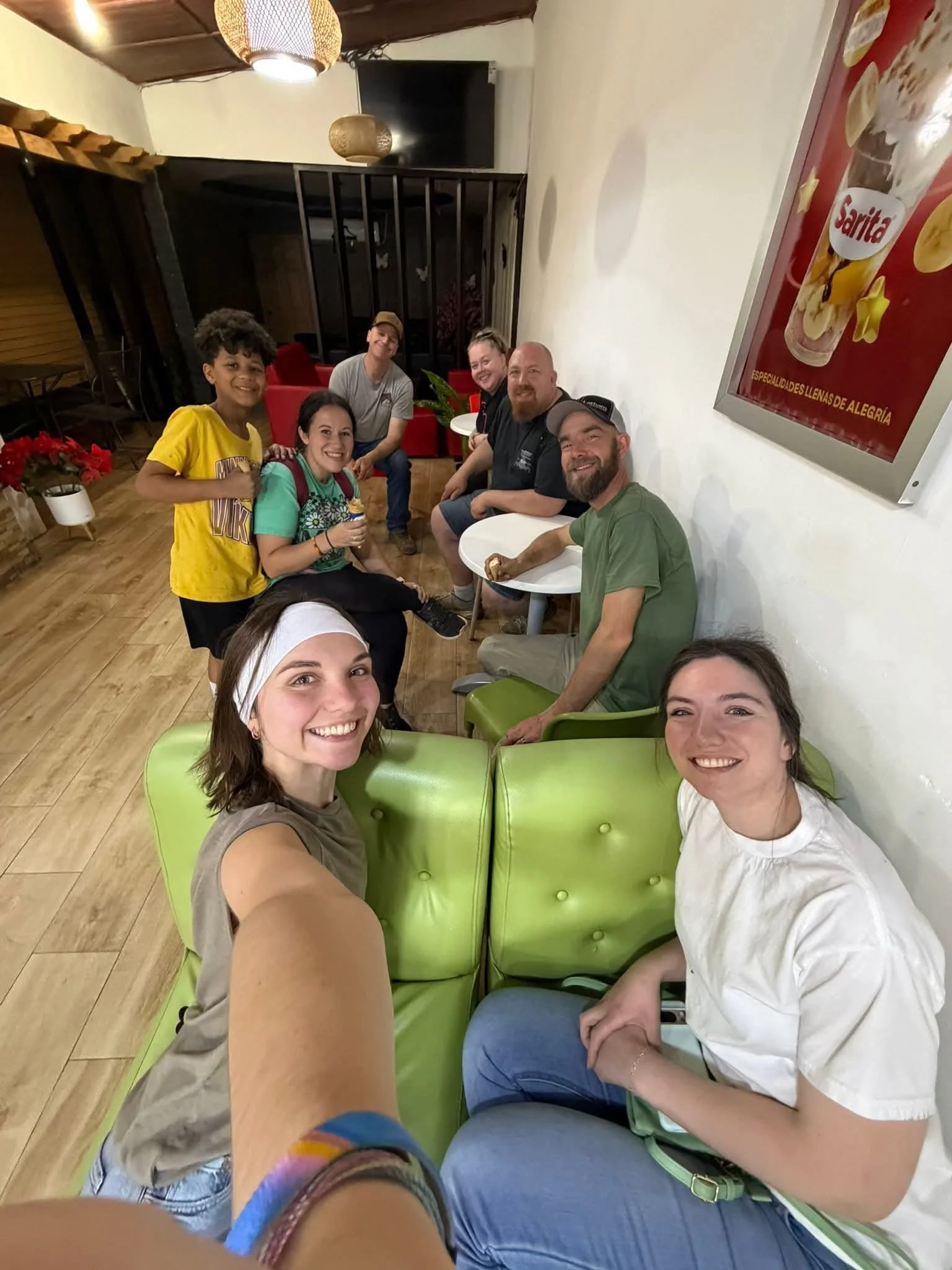 A group of six smiling people sitting and standing in a cozy cafe or restaurant. They are casually dressed, with some holding food or drinks. The setting features wooden flooring, green seating, white walls, and a framed poster on the wall.