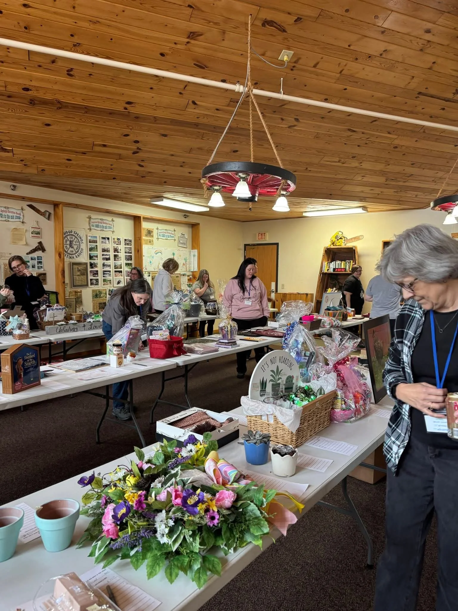 Indoor event with tables displaying various items including flowers, gift baskets, and decorative objects. Several people are browsing and arranging items in a room with wooden ceiling panels and informational posters on the walls.