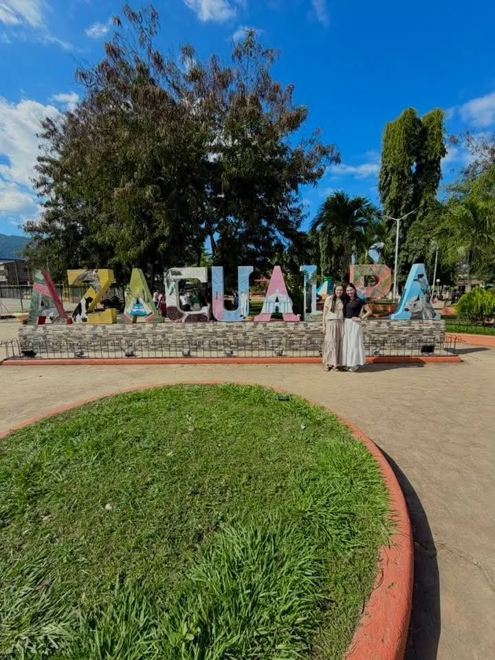 Two women standing in front of a colorful sign that spells out 'Aguascalientes' in a park with green trees, a clear blue sky, and a grass patch in the foreground.