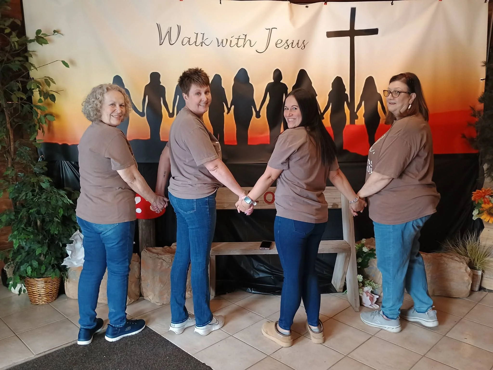 Four women are holding hands in a circle, standing in front of a backdrop with silhouettes of people holding hands and a cross, with the text "Walk with Jesus". They are indoors with plants on either side.
