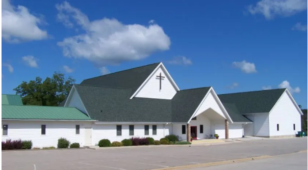 White church with a cross on its steeple, green roof, blue sky with clouds, and a parking lot in front.