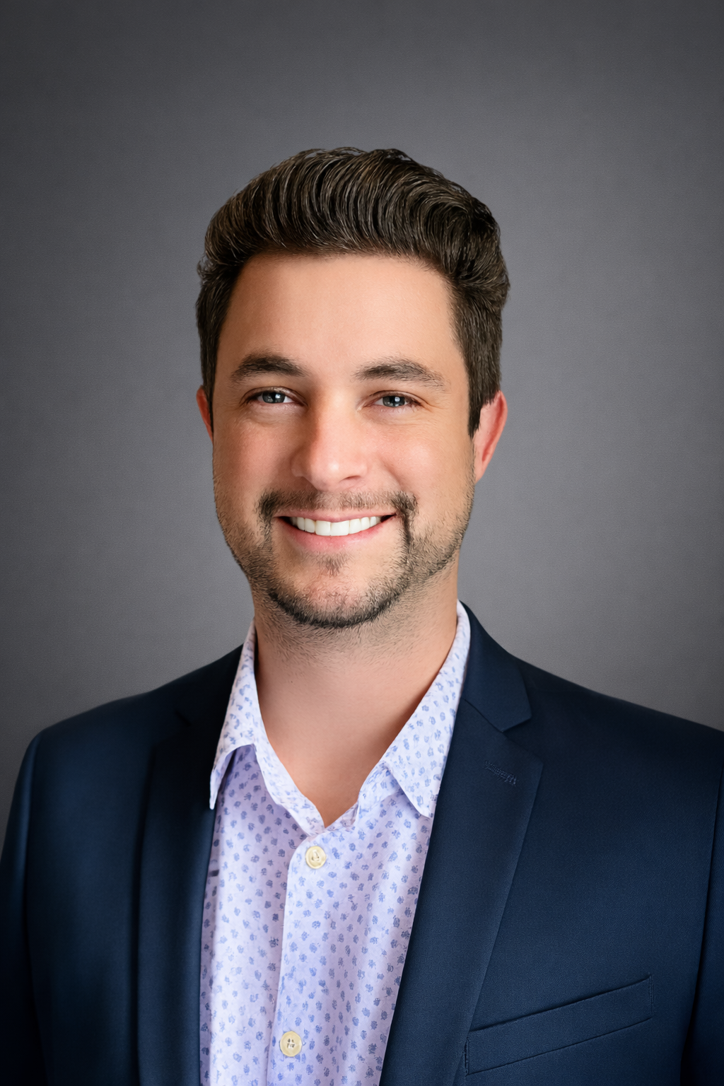 Headshot of a smiling man with dark hair, wearing a dark suit jacket and a light patterned dress shirt against a gray background.