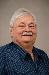 Portrait of an elderly man with gray hair and a mustache, smiling, wearing a blue striped shirt.