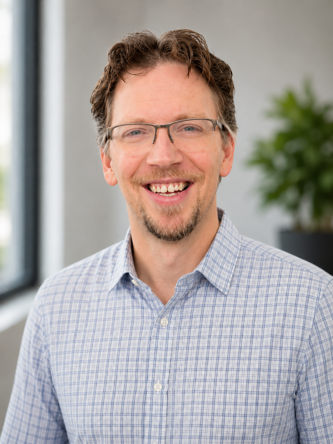 Photograph of a smiling man with glasses, light blue checkered shirt, light brown hair, and a beard, indoors with a window and a green plant in the background.