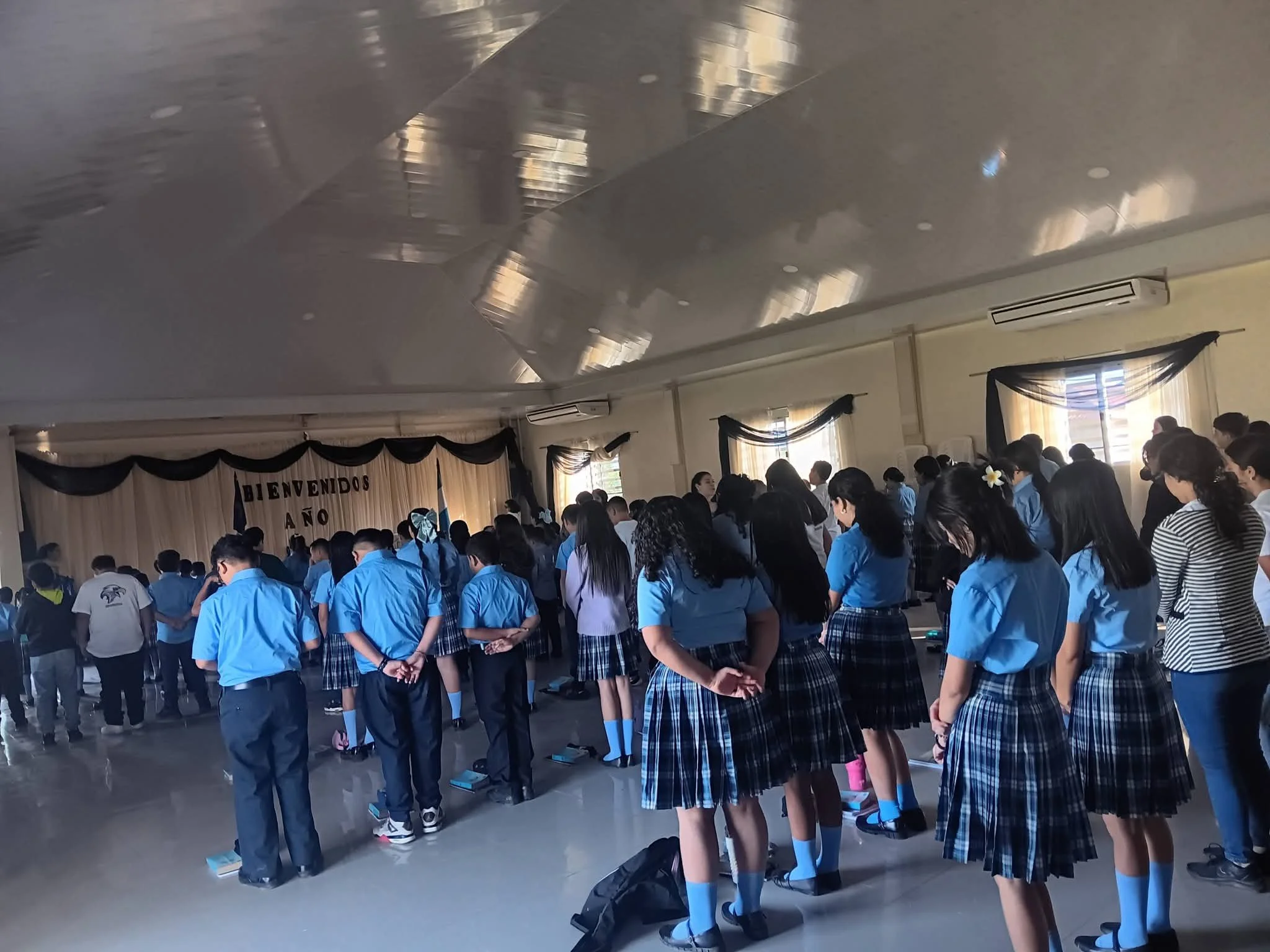 Students standing in a line in a school gymnasium with uniforms, facing forward, preparing for an event or ceremony.