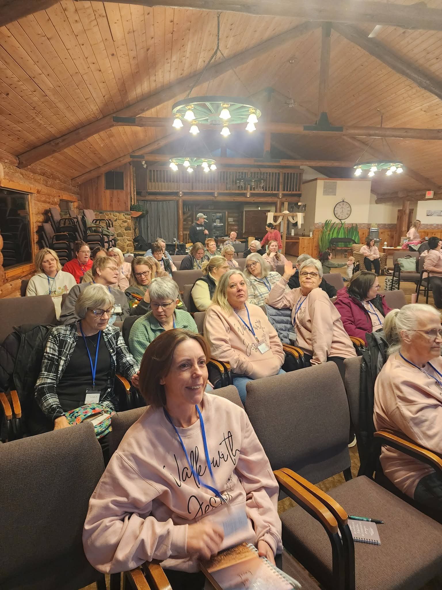 A large group of women seated in a wood-paneled room with log cabin style decor. Some are looking at the camera, others talking, with a few taking notes. The room has rustic chandeliers and a staircase in the background.