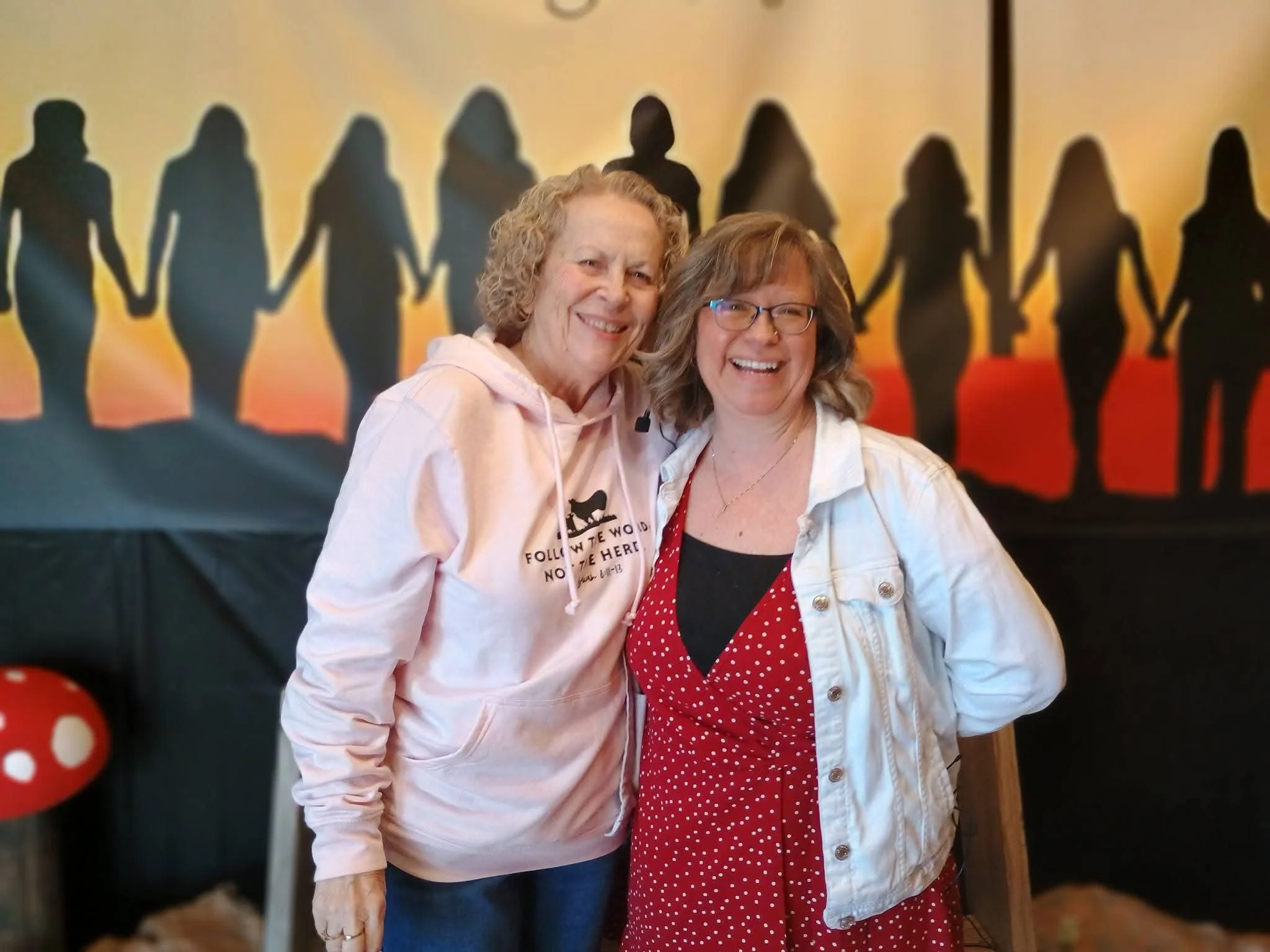 Two women smiling and standing close together, with shadow silhouettes of people holding hands on a colorful background behind them.