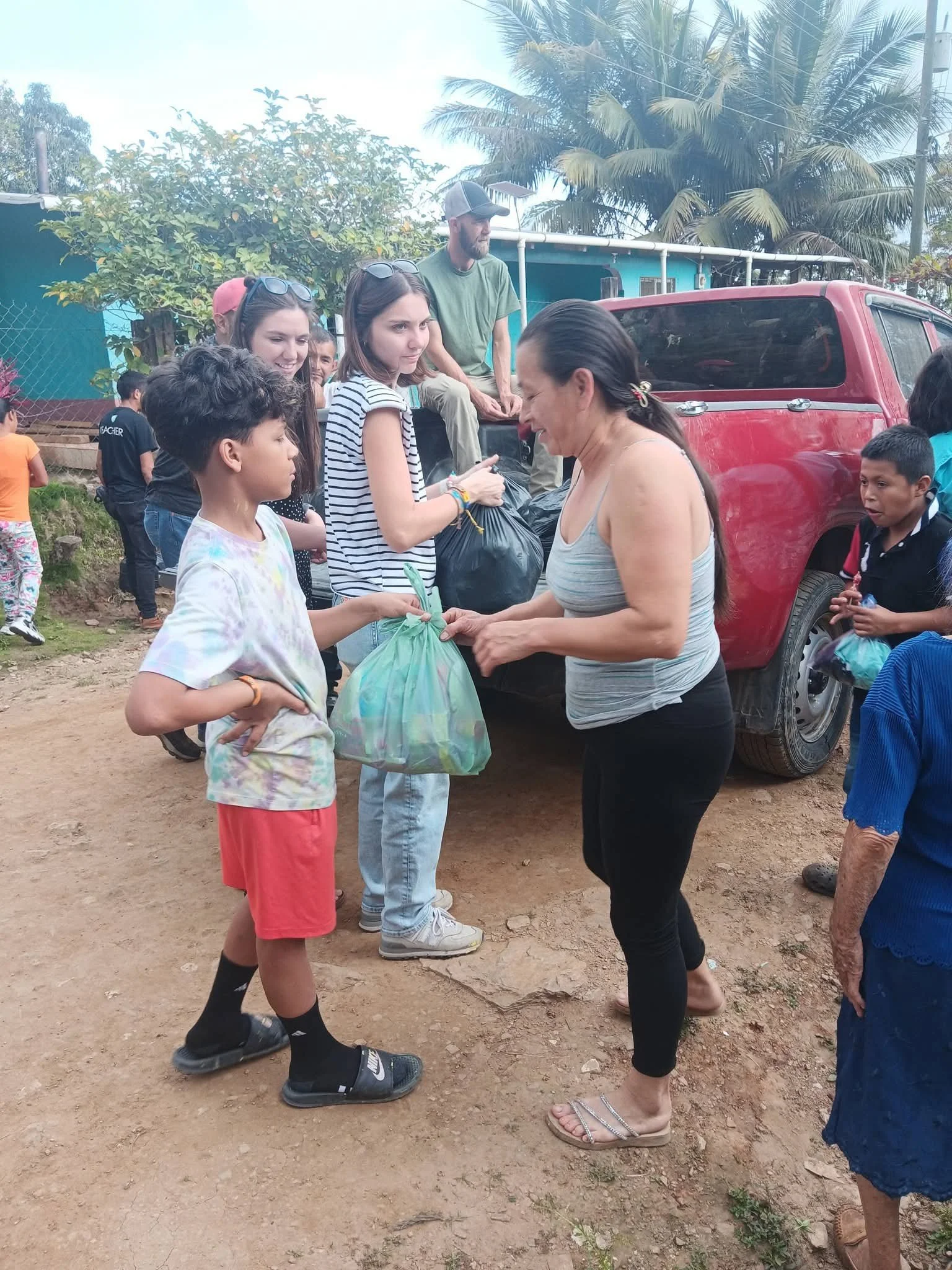 Group of people, including children, women, and men, standing and interacting outside near a red vehicle with tropical trees in the background. One woman appears to be giving something to a young boy.