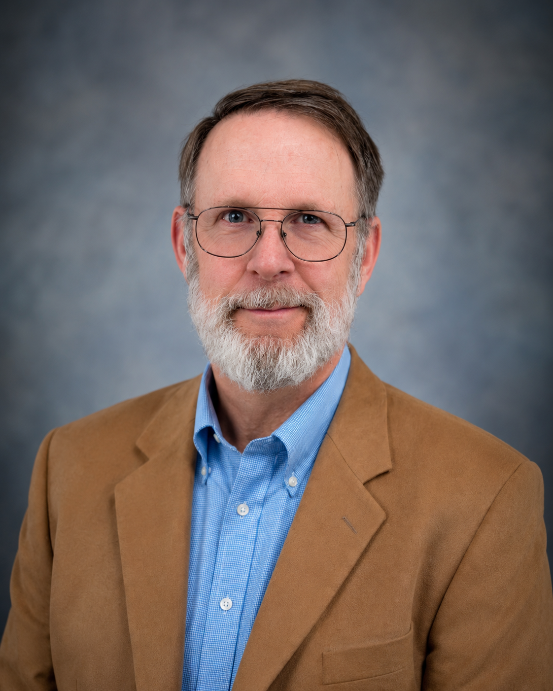 A professional portrait of a senior man with glasses, a gray beard, wearing a tan blazer and a blue collared shirt against a gray background.