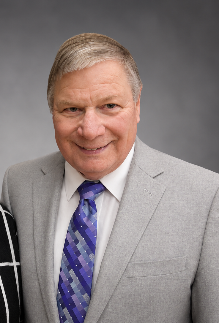 A smiling middle-aged man with light blond hair dressed in a light gray suit, white shirt, and a purple patterned tie, standing against a neutral gray background.