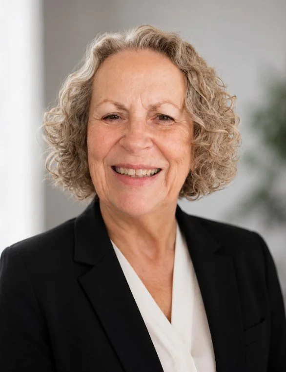An older woman with curly gray hair smiling, wearing a black blazer and white top, standing indoors with a blurred background.