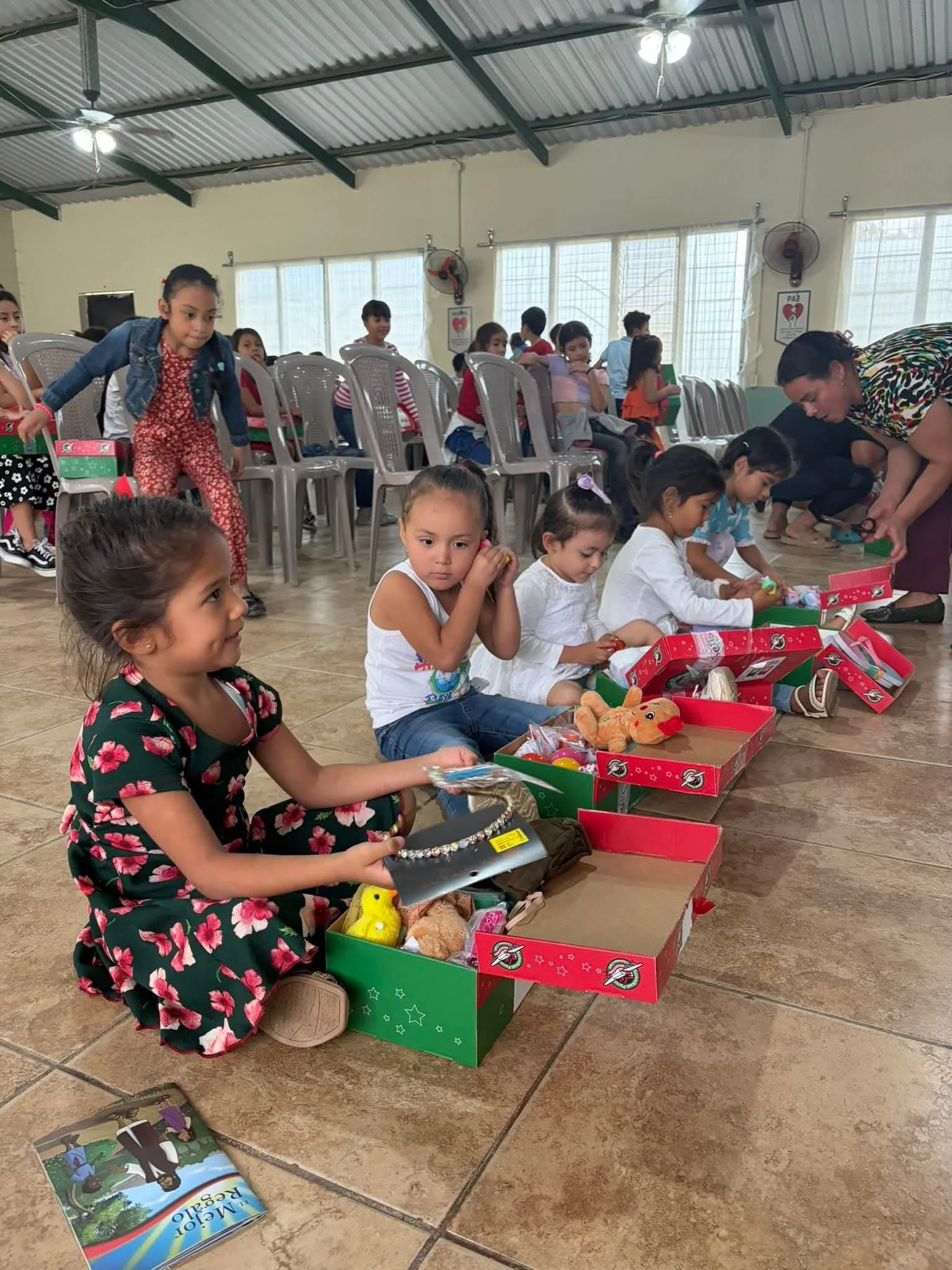 Children sitting on the floor and in chairs, opening Christmas presents inside a community center or hall.