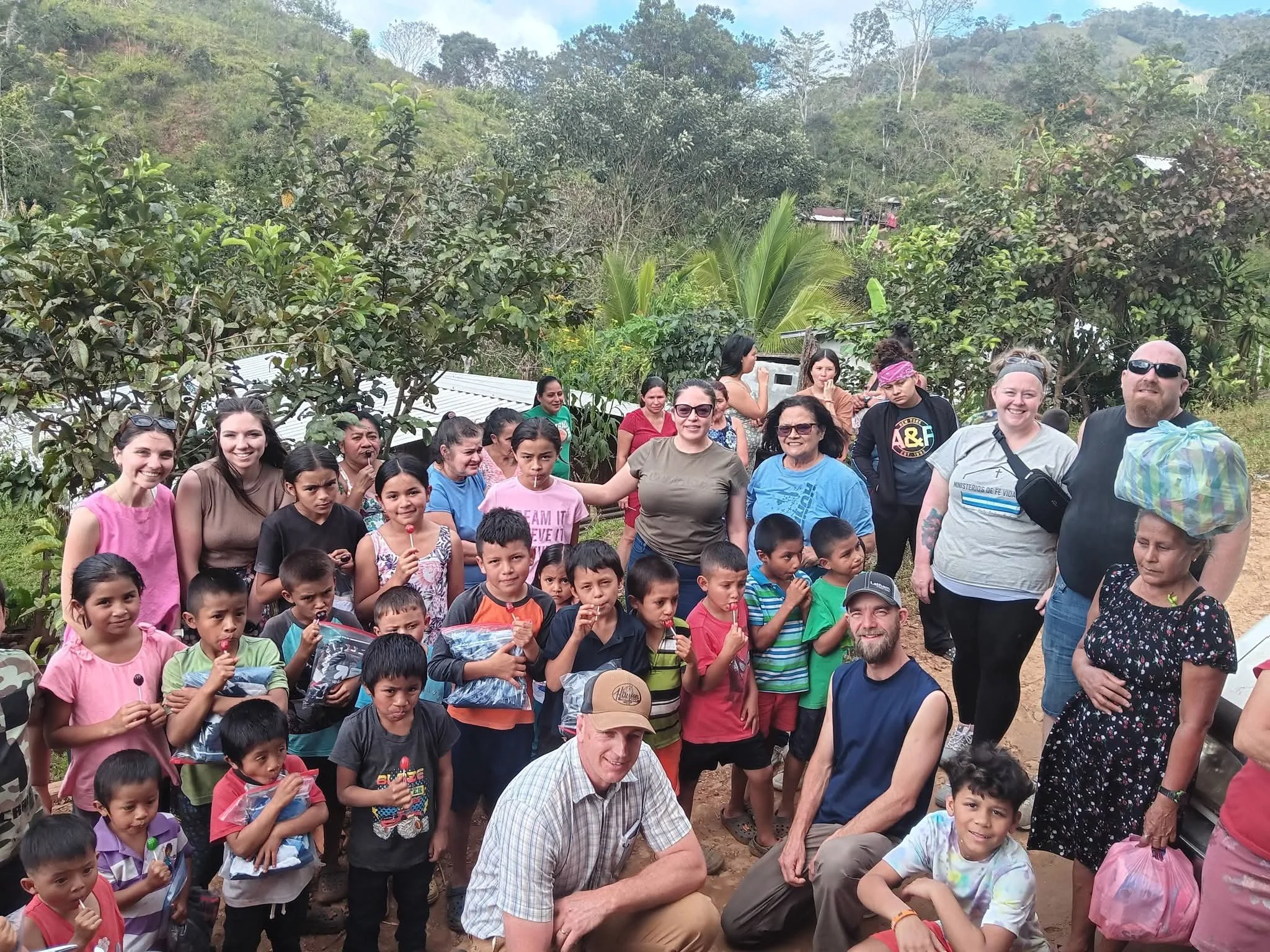 A large group of people, including children and adults, gathered outdoors in a rural area with trees and mountains in the background. Some are smiling and holding items, and there is a man in a striped shirt in the foreground.