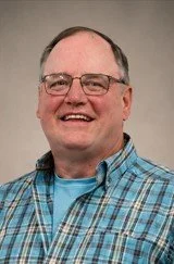 A smiling middle-aged man wearing glasses, a blue plaid shirt, and a light blue T-shirt standing against a plain background.