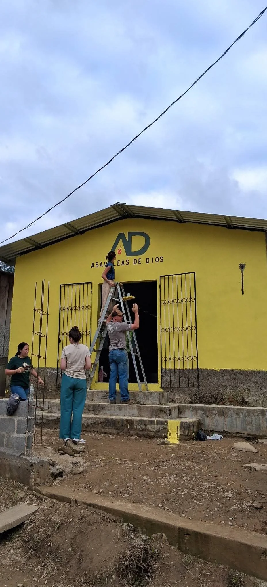 People painting a yellow church with the name 'Asamblea de Dios' and the initials 'AD' on the front.