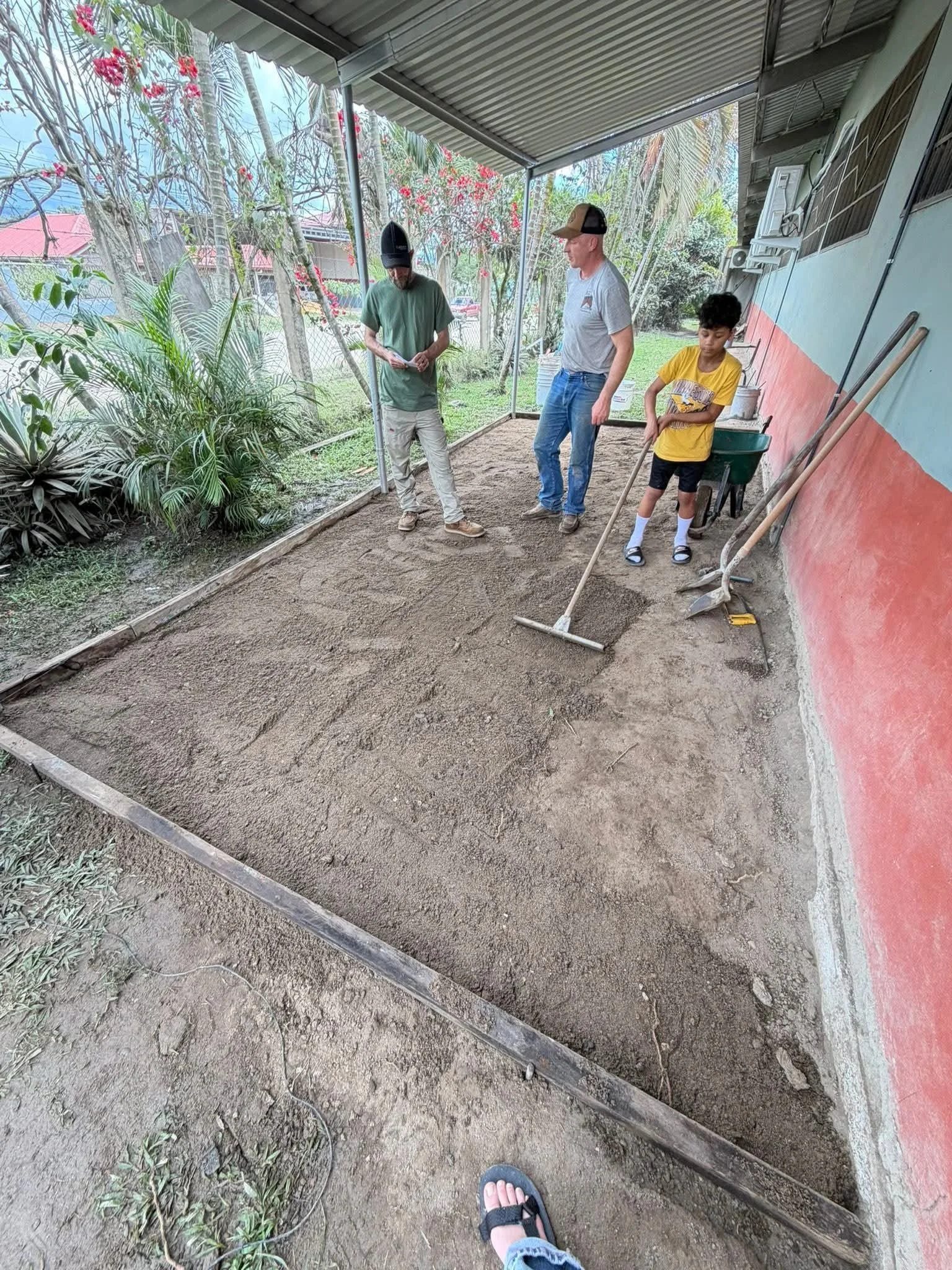 A group of three individuals, two adult males and a young boy, are standing in a partially enclosed outdoor area with a dirt floor. They are observing the ground, which appears to be prepared for planting or gardening, surrounded by plants and trees,