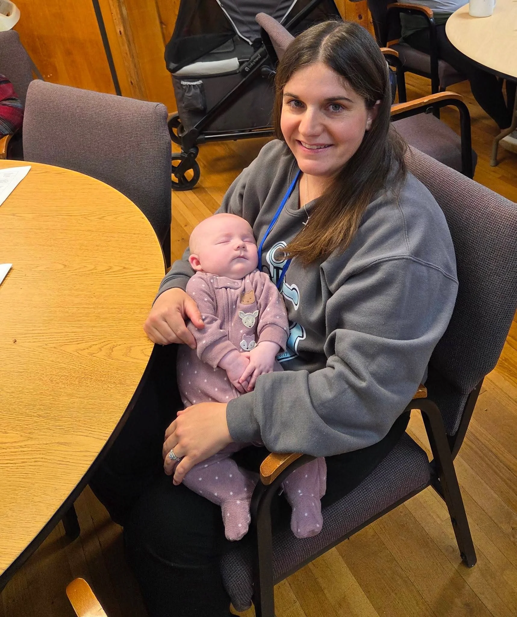 A woman with long brown hair smiling at the camera, holding a sleeping baby dressed in pink with white polka dots in her arms, sitting in a restaurant or cafe with wooden walls and round tables.