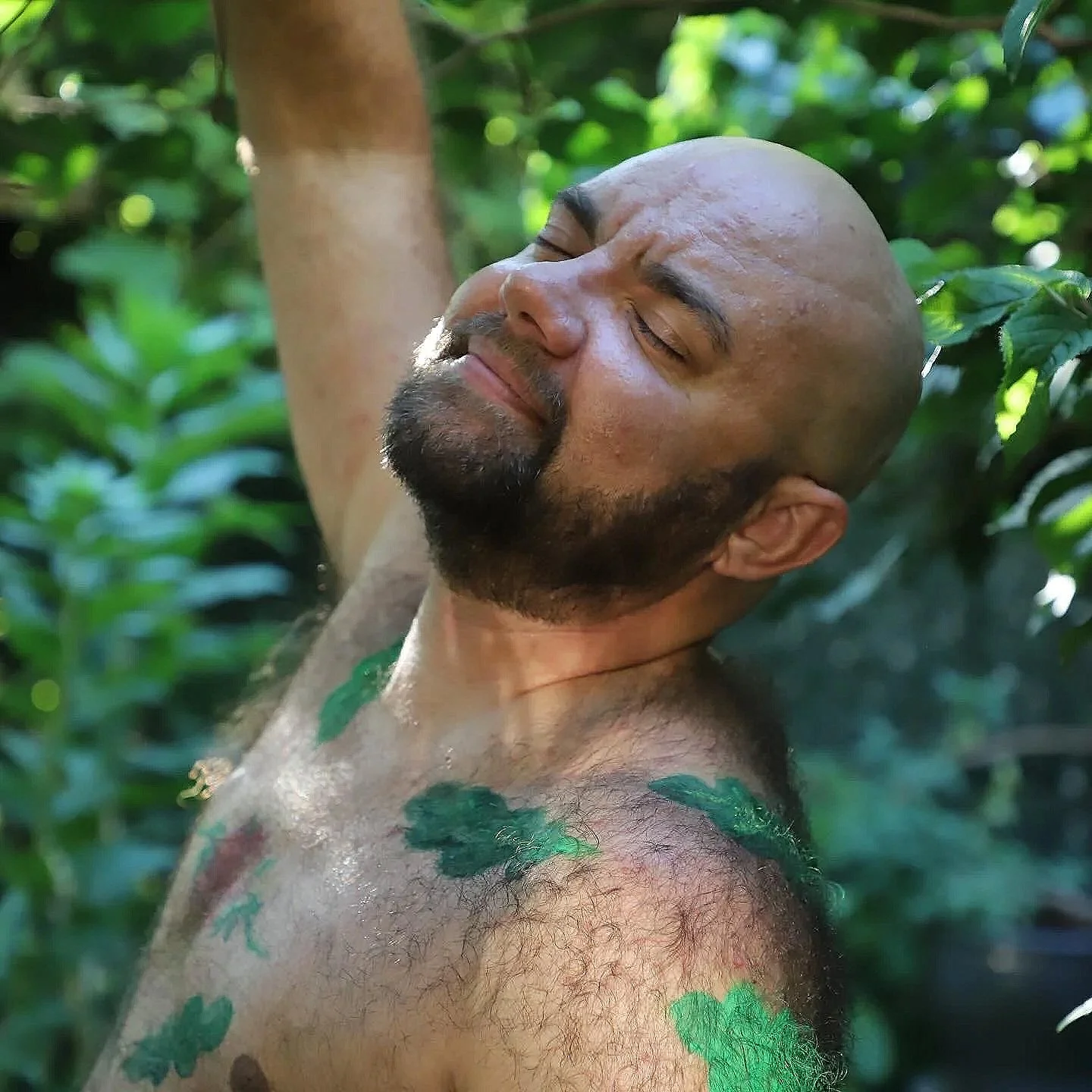 a white bald person with a beard, eyes closed, smiling surrouned by green leaves
