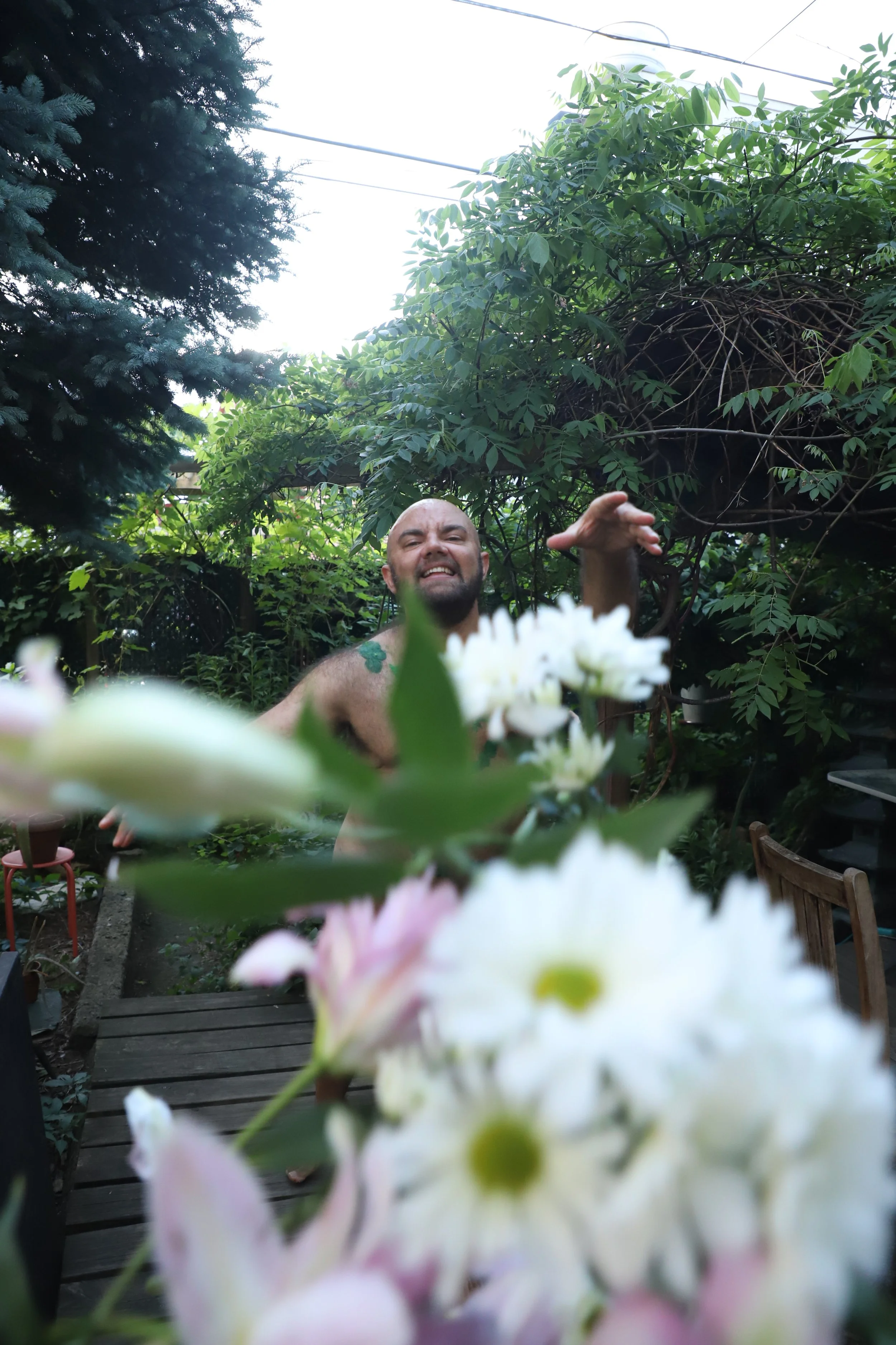 A shirtless white person with a beard, smiling and reaching out, standing outdoors among green plants and trees, with a wooden table and chairs nearby. Blurred white and pink flowers are in the foreground.
