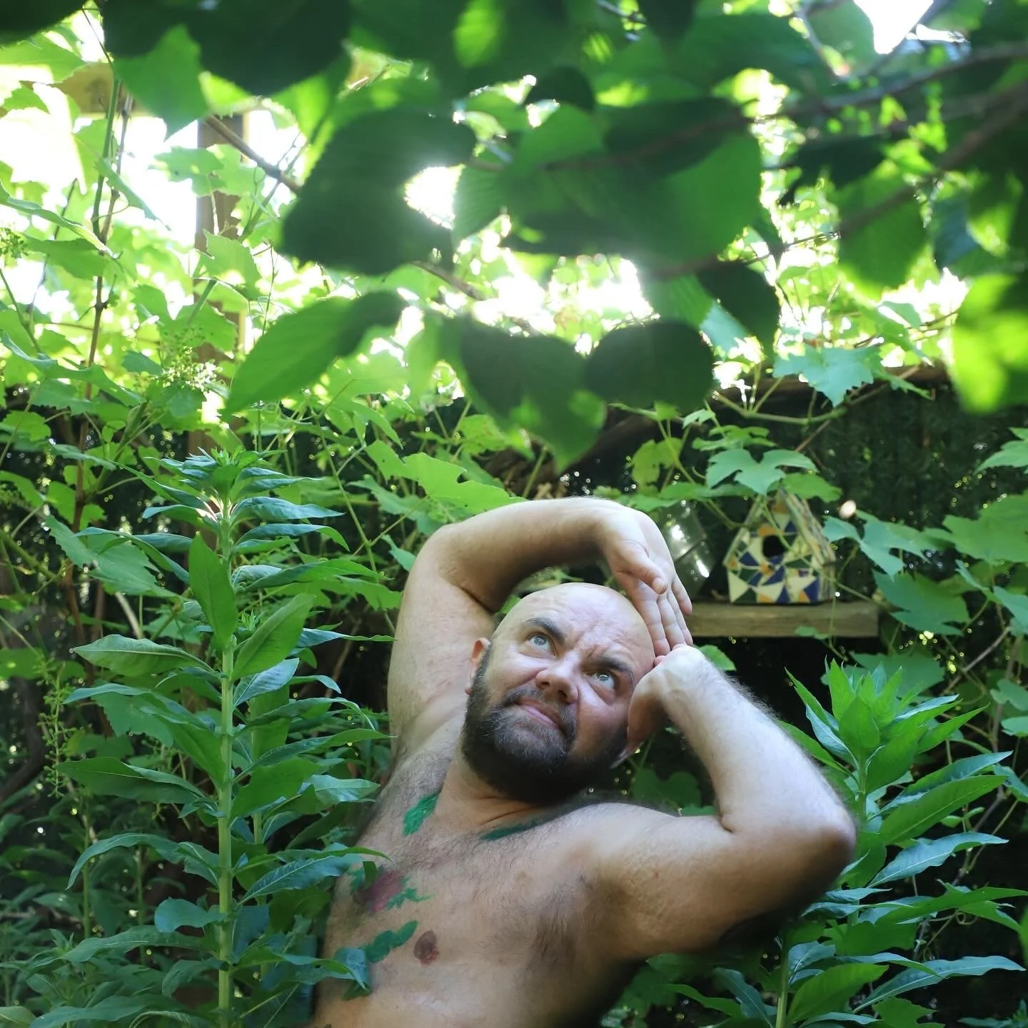 A shirtless white person with a beard and tattoos poses among lush green plants and leaves, looking upward with one arm over his head in a natural outdoor setting.