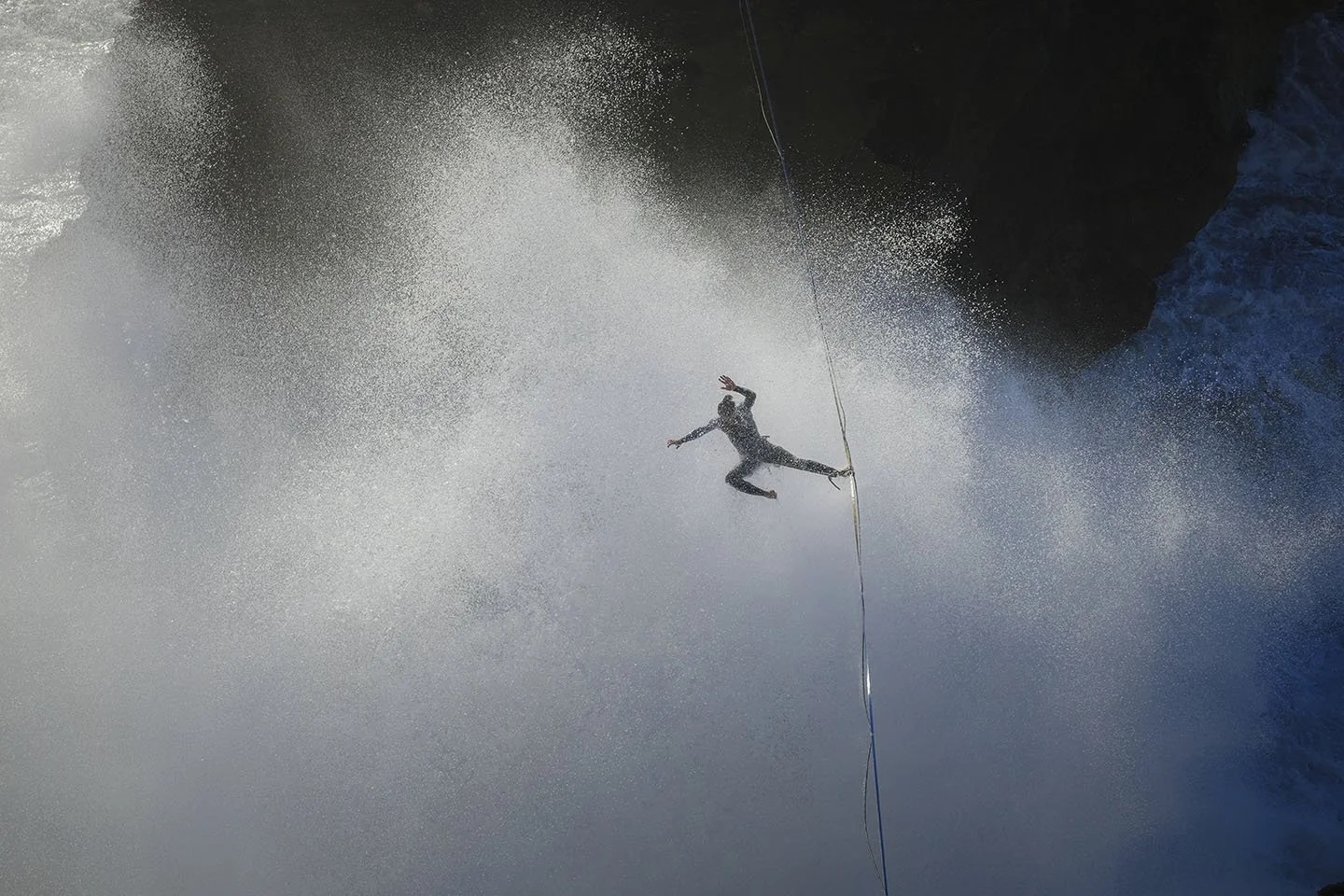 Person mid-air, holding a rope, near water spray, possibly rock climbing or rappelling near a waterfall.