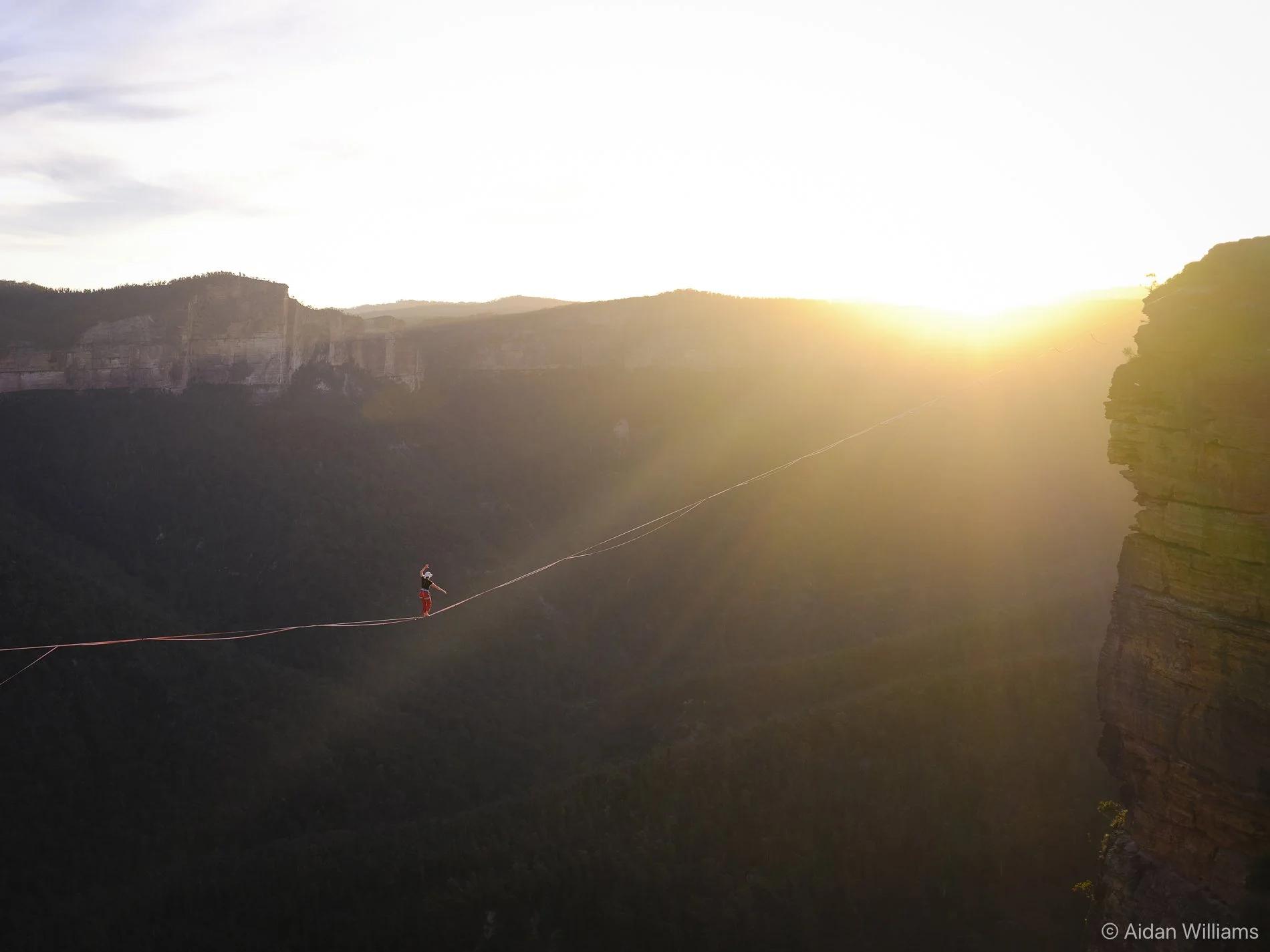 Person walking on a tightrope high above a deep canyon during sunset with cliffs in the background.