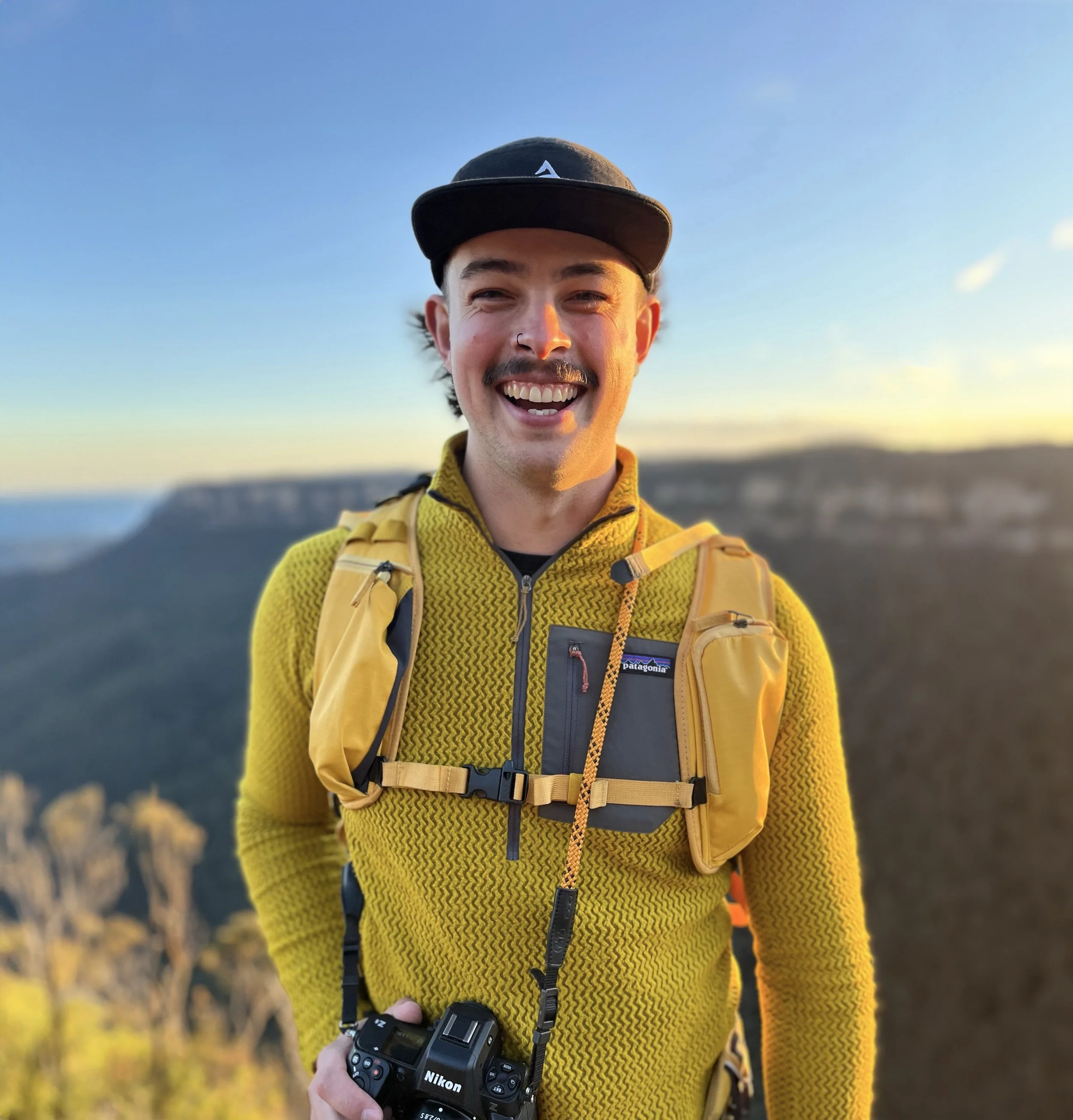 A smiling man in a yellow sweater and black cap outdoors during sunset, holding a Nikon camera, with a blurred scenic landscape in the background.