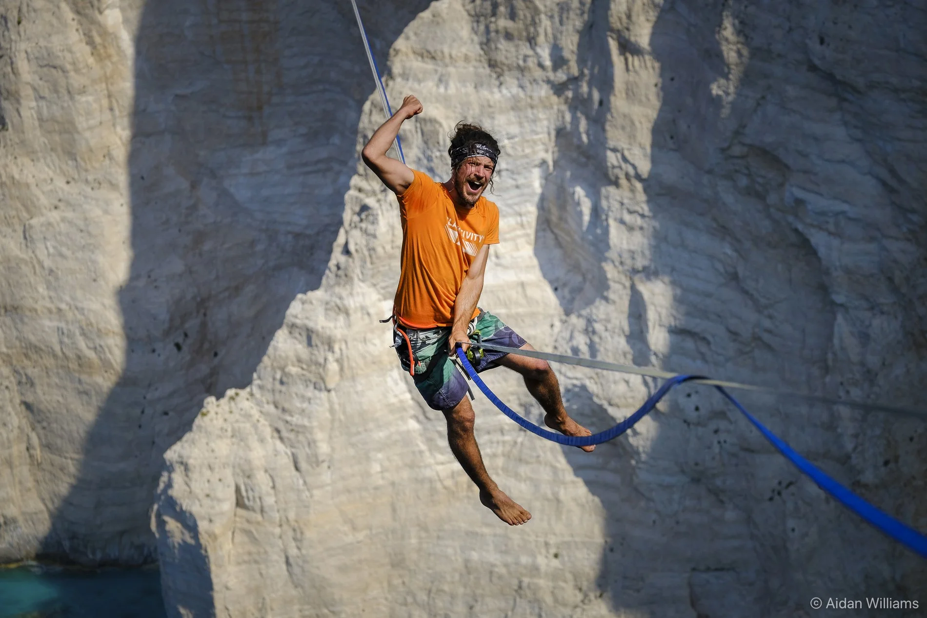 A man with long hair, wearing an orange shirt and multicolored shorts, is mid-air on a slackline set against white rocky cliffs, appearing to be enjoying an outdoor adventure near water.
