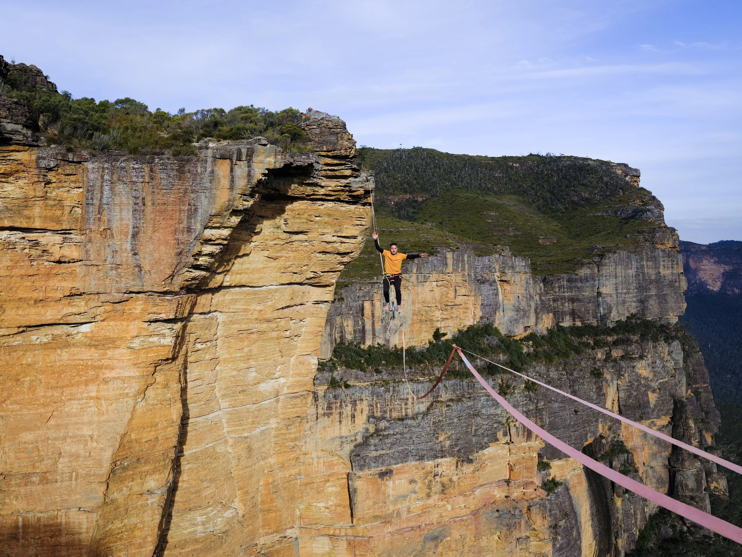 A person on a zip line crossing a deep canyon with rugged rock cliffs and green vegetation, under a partly cloudy sky.