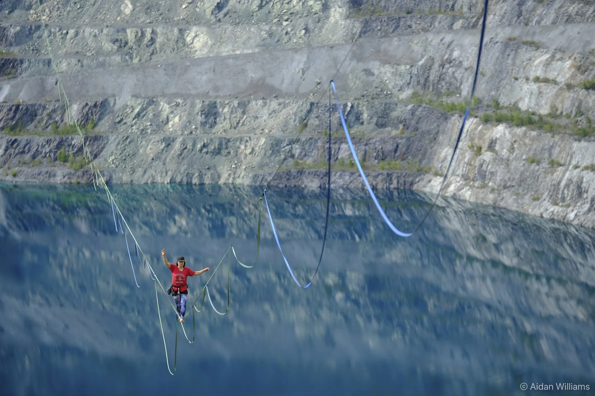 A person balancing on a tightrope stretched over a body of water with steep rocky cliffs in the background.