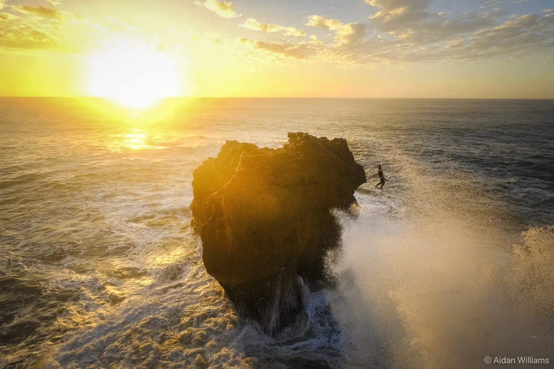 A person wakesurfing on the ocean near a large rock at sunset, with the sun low on the horizon and waves crashing around.