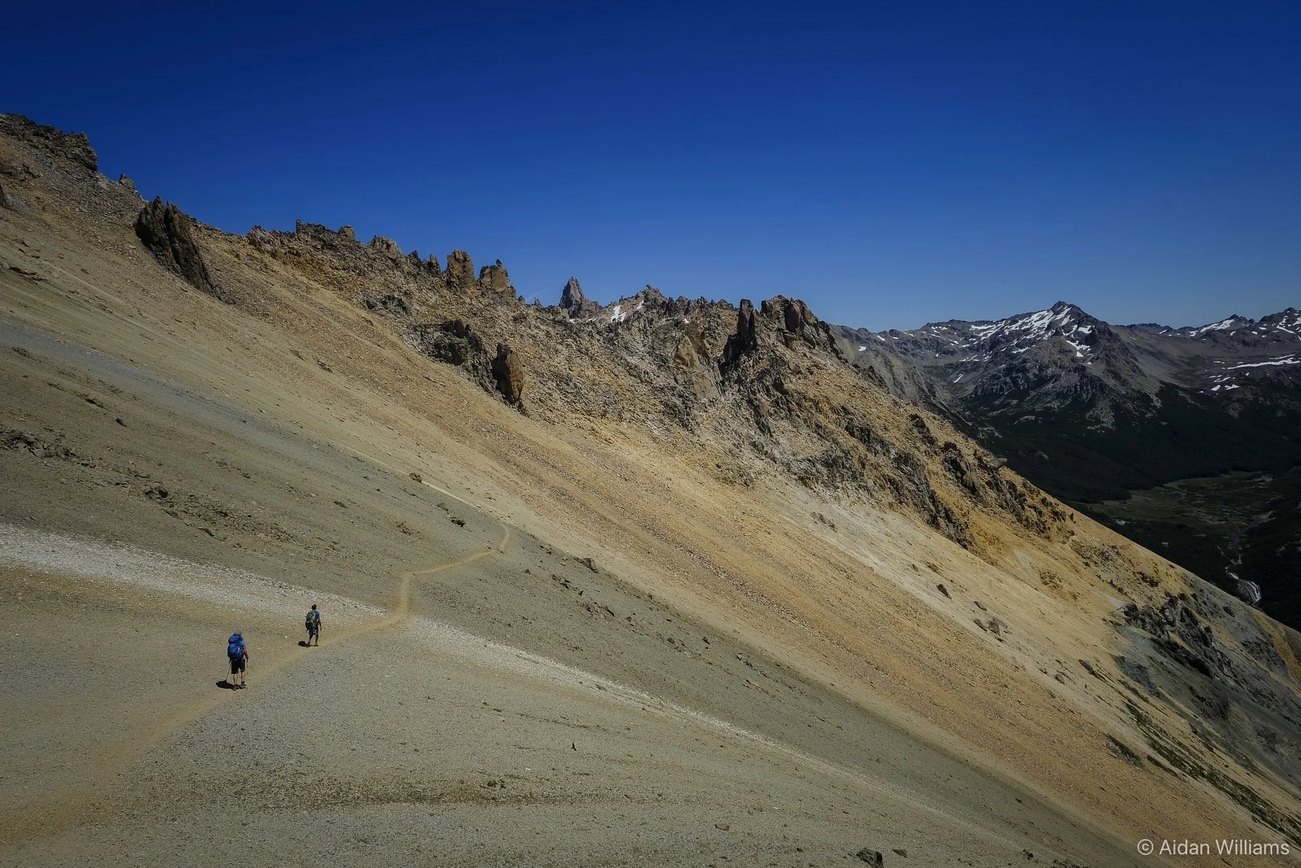 Two hikers with backpacks walking along a winding trail through a barren, rocky mountain landscape under a clear blue sky.