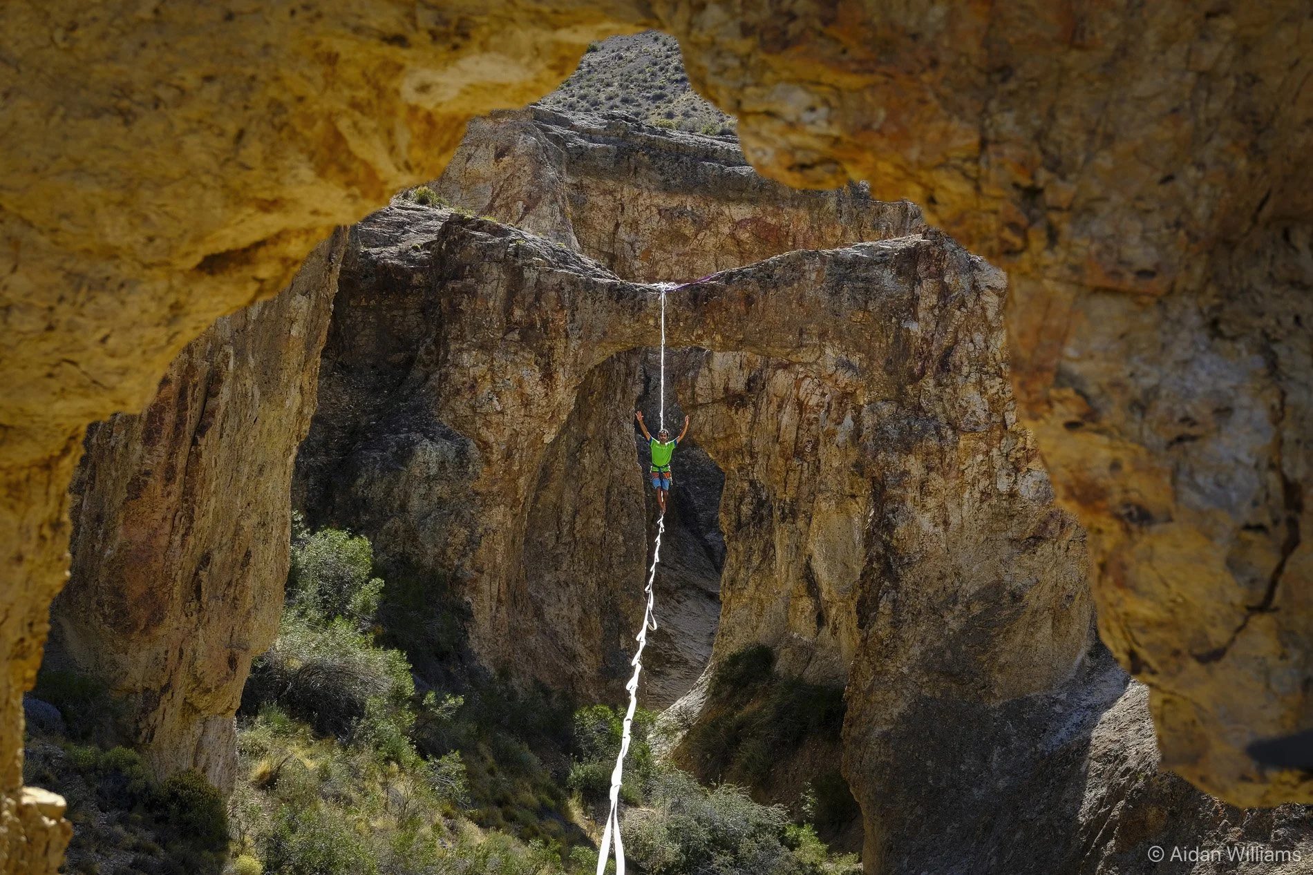 A person in a green shirt and blue shorts is slacklining between rock formations in a canyon, with arms raised and a safety harness attached to the slackline.