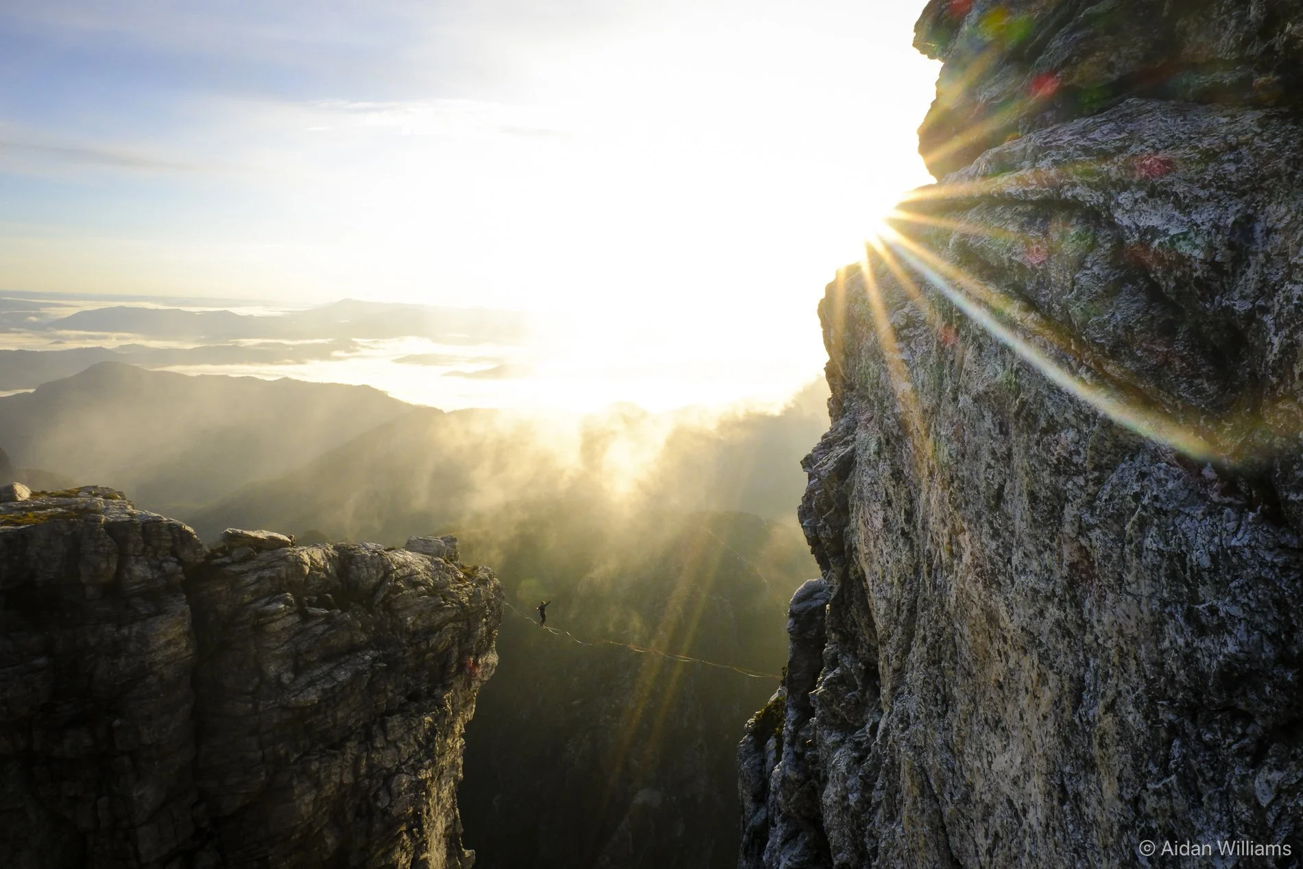 A mountain climber on a rope between two tall rocky cliffs during sunrise with the sun shining over the horizon and casting rays of light.