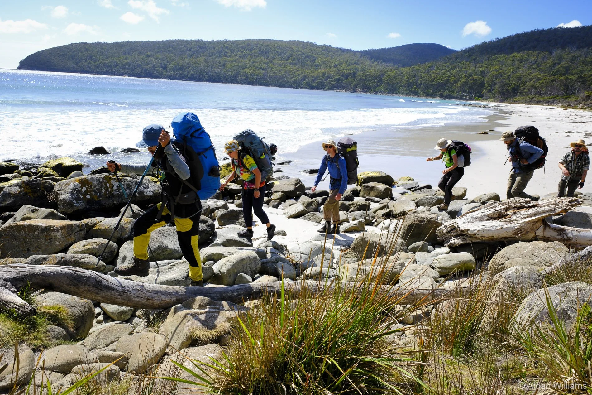 A group of hikers crossing a rocky beach near the ocean, with a forested hill in the background, under a partly cloudy sky.