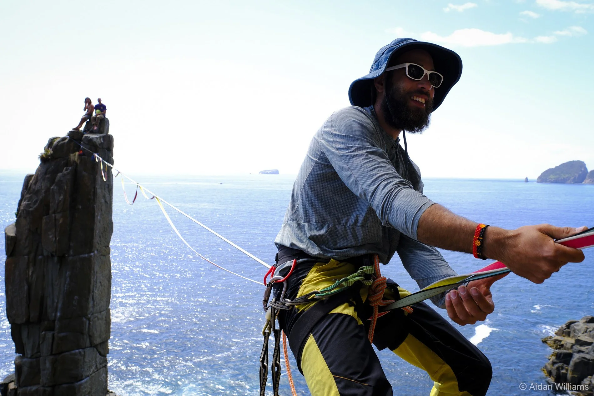 A man in outdoor gear, including a hat, sunglasses, and a harness, is standing near the ocean, holding a tablet or phone, with a rocky cliff in the background. Two people are sitting on top of the cliff.