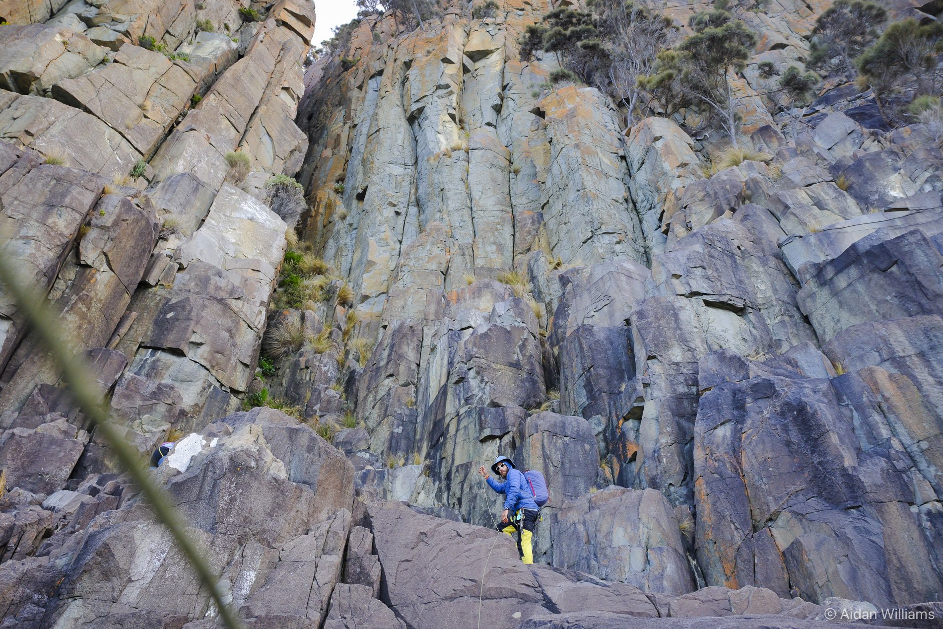 A person wearing a blue jacket, yellow pants, sunglasses, and a helmet is mountain climbing on large rocks and boulders at the base of a steep, colorful rocky cliff with sparse patches of vegetation.