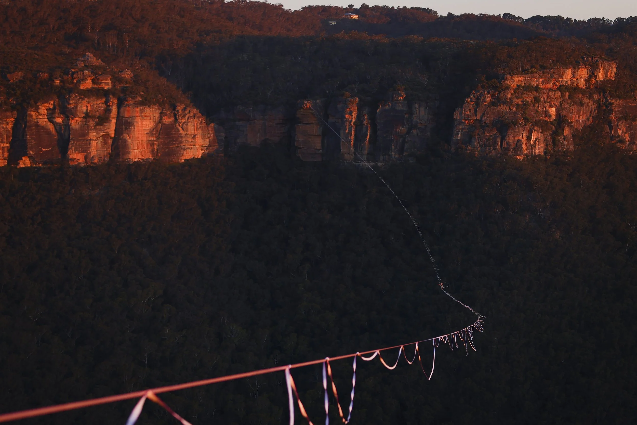 A long suspension bridge extends across a deep valley surrounded by forested hills, with a rocky cliff face illuminated by warm orange light in the background.