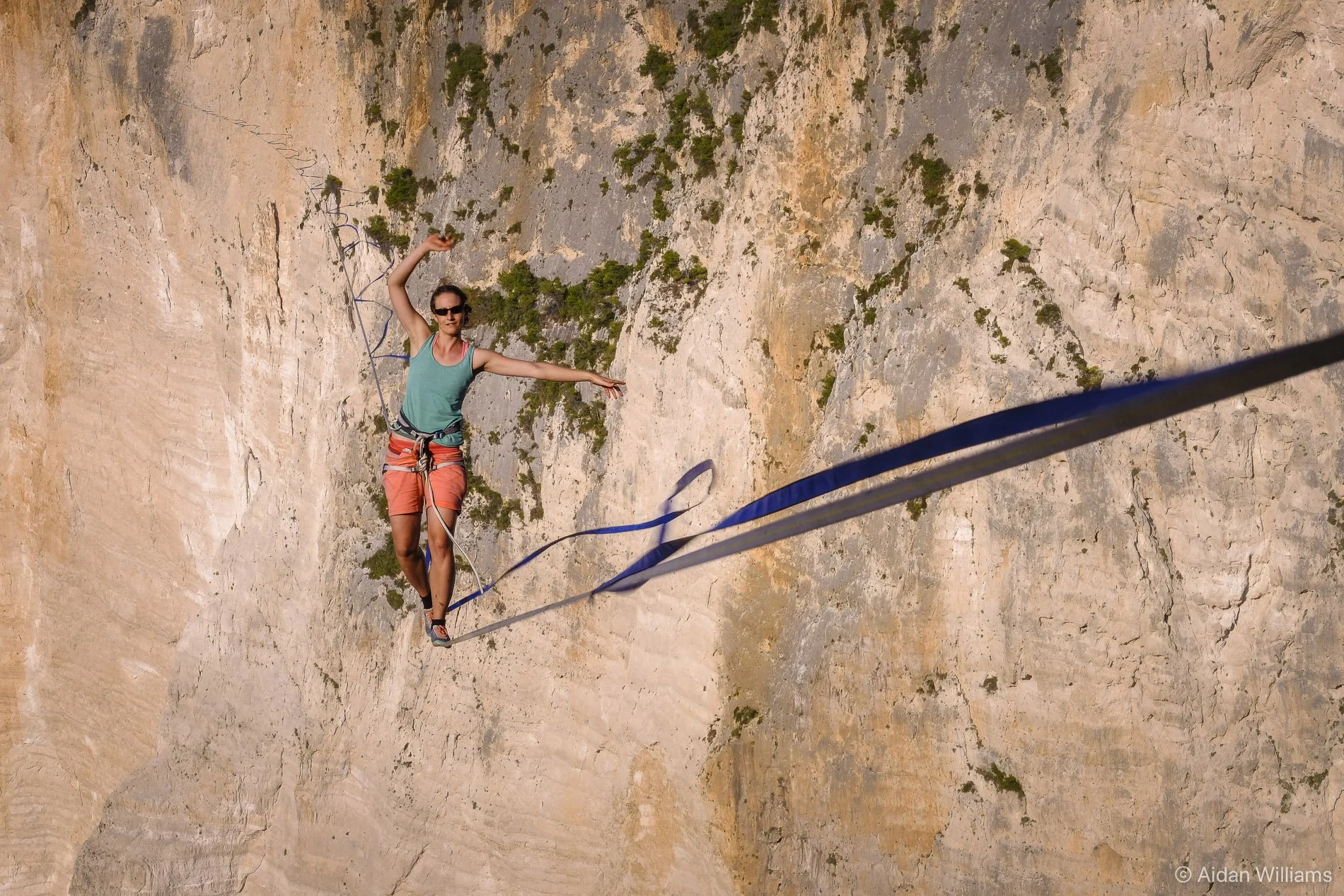 A woman slacklining on a narrow line attached between a rock face and a rope, with one arm extended for balance, in an outdoor setting with steep rocky cliffs and sparse vegetation.