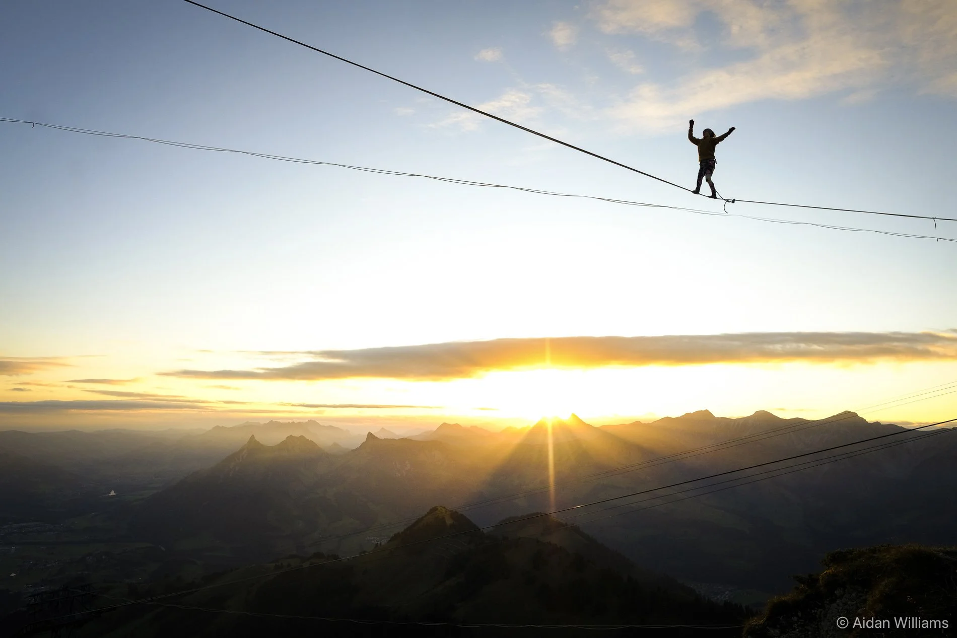Person walking on a tightrope high above a mountainous landscape at sunset.