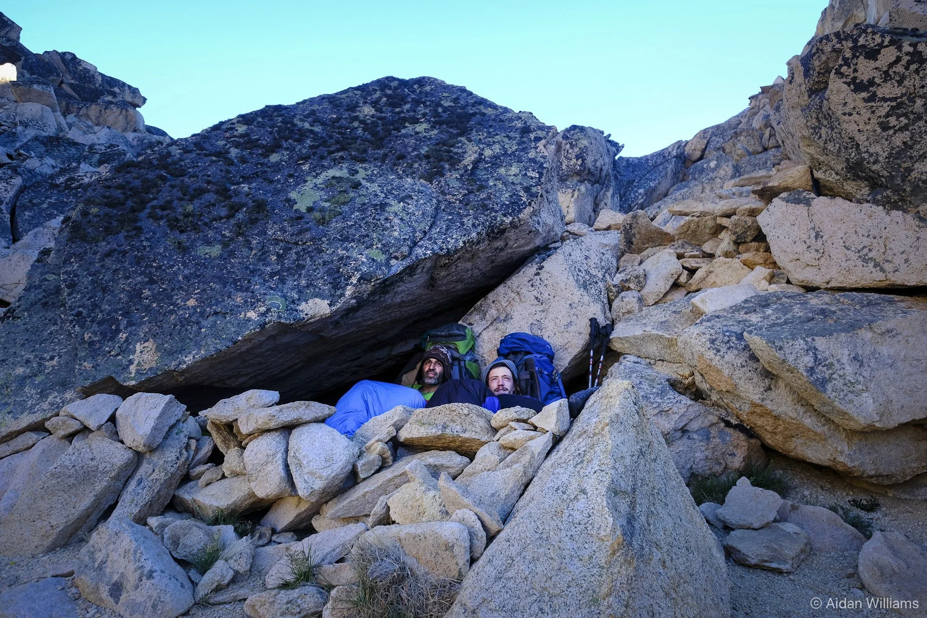 Two hikers resting beneath a large rock formation on a mountain trail.